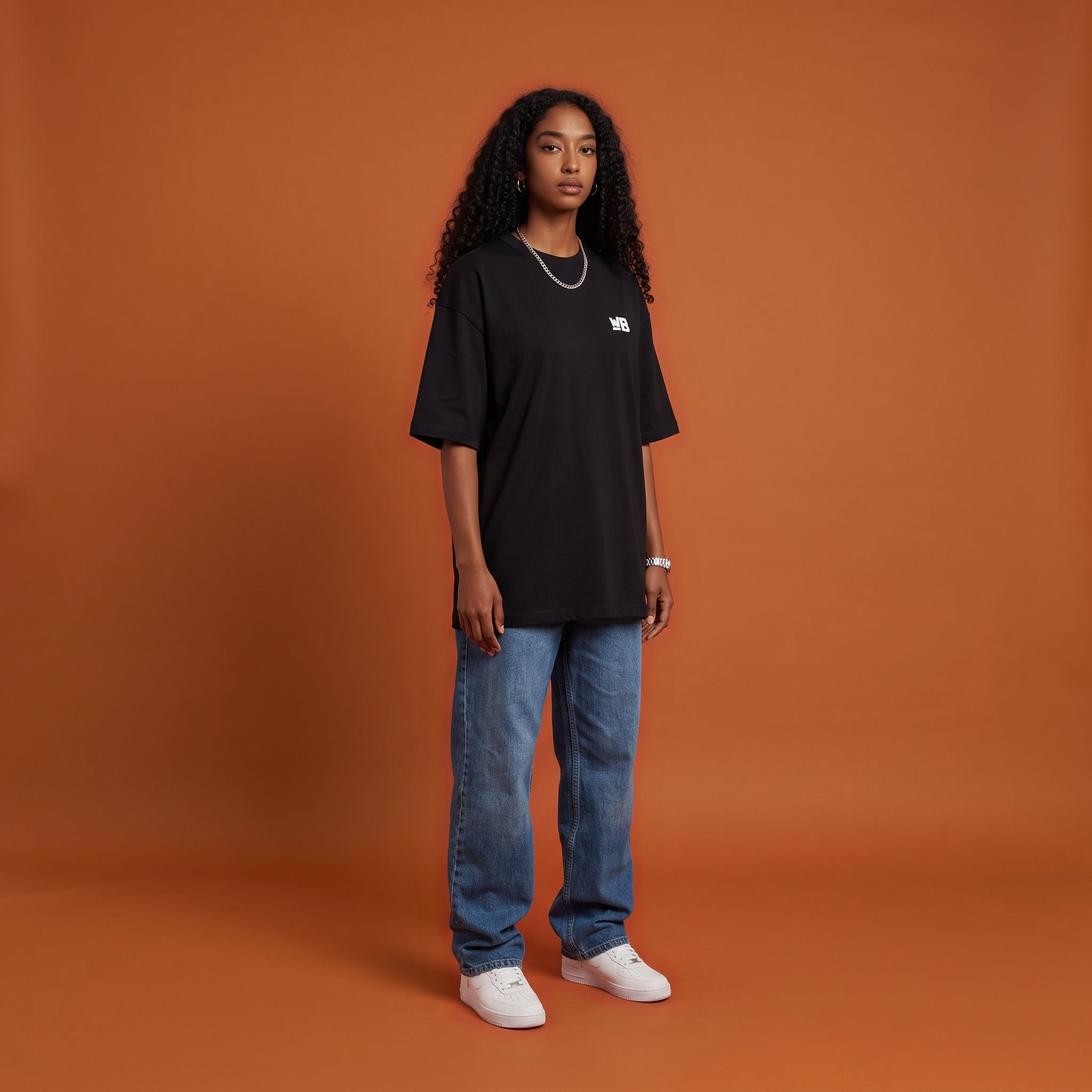 Blackness Sankofa Queen Tee, black, worn by a model — front view with WB chest logo, burnt-orange terracotta studio backdrop