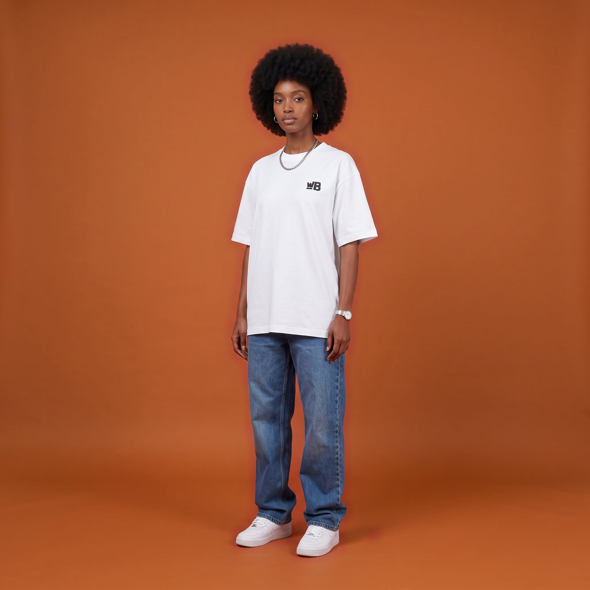 Blackness Sankofa Queen Tee, white, worn by a model — front view with WB chest logo, burnt-orange terracotta studio backdrop