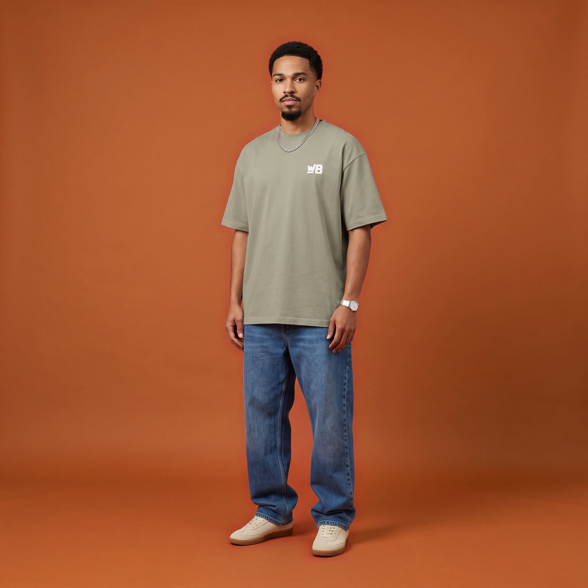 Blackness Sankofa King Tee, brown khaki, worn by a model — front view with WB chest logo, burnt-orange terracotta studio backdrop
