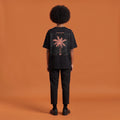 Blackness Libation Tee, black, worn by a model — back view, palm tree Libation print, terracotta-orange studio backdrop