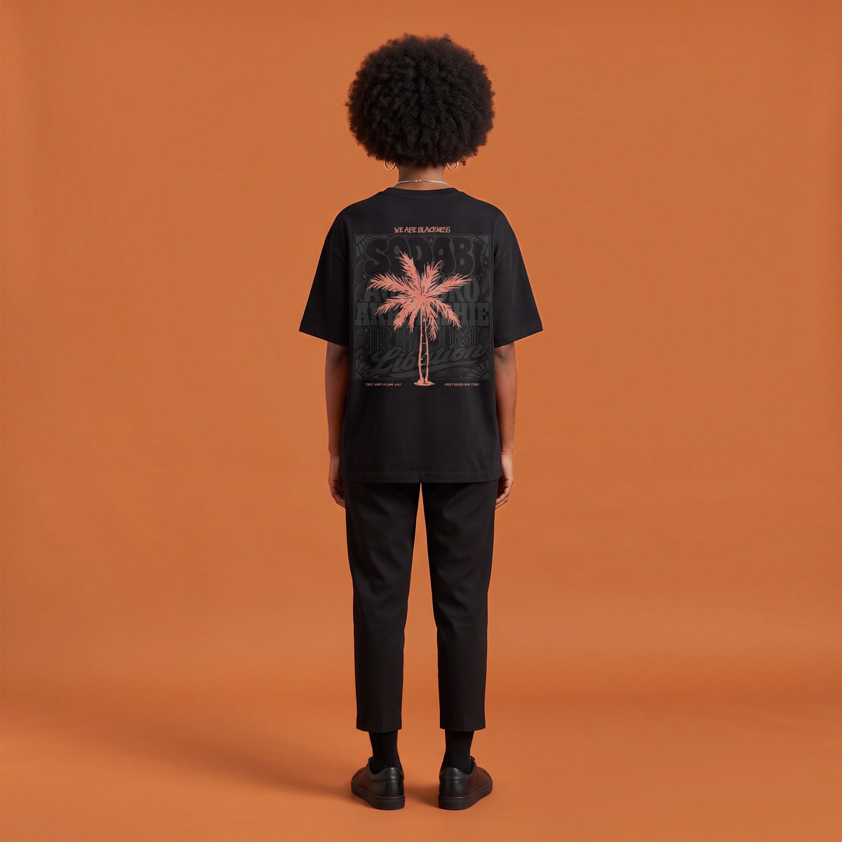 Blackness Libation Tee, black, worn by a model — back view, palm tree Libation print, terracotta-orange studio backdrop