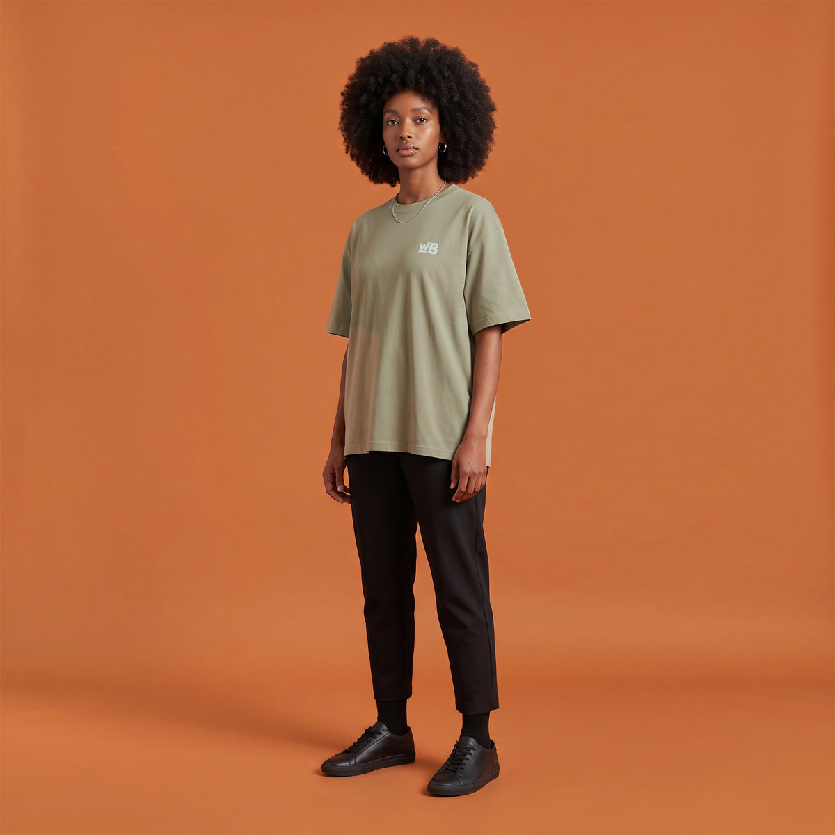 Blackness Libation Tee, brown khaki, worn by a model — front view with WB chest logo, terracotta-orange studio backdrop
