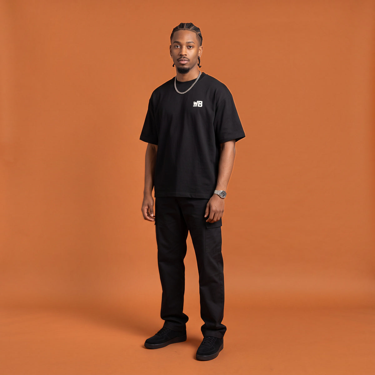 Blackness Mawon Tee, black, worn by a model — front view with WB chest logo, terracotta-orange studio backdrop