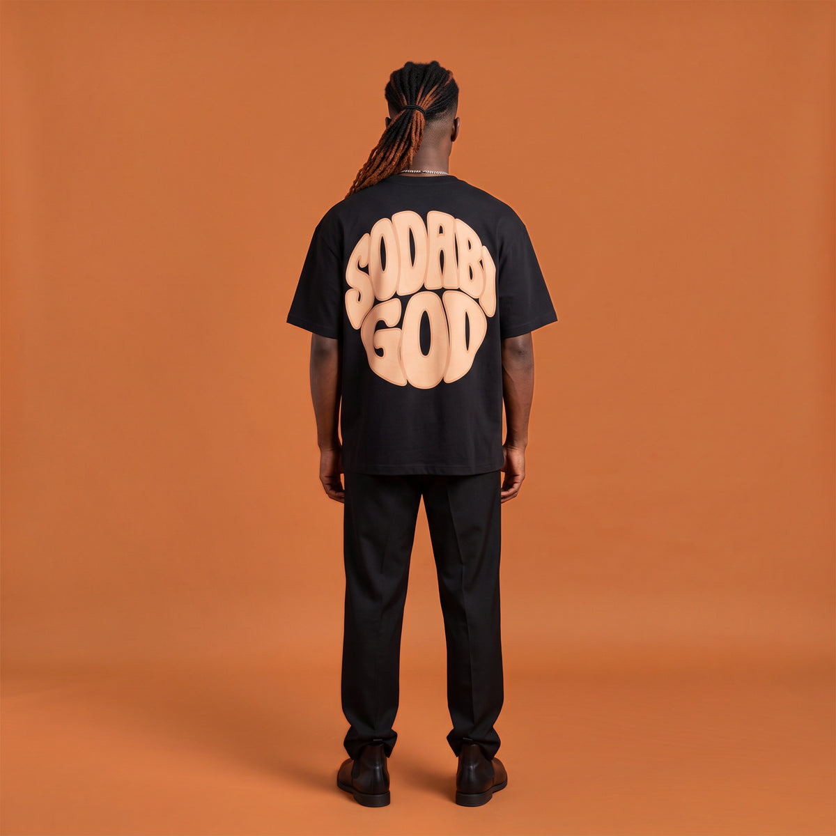 Blackness Sodabi Tee, black, worn by a model — back view, SODABI GOD bubble lettering print, terracotta-orange studio backdrop