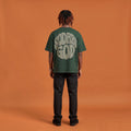 Blackness Sodabi Tee, forest green, worn by a model — back view, SODABI GOD bubble lettering print, terracotta-orange studio backdrop