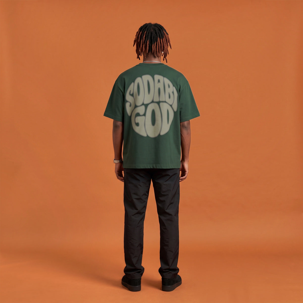 Blackness Sodabi Tee, forest green, worn by a model — back view, SODABI GOD bubble lettering print, terracotta-orange studio backdrop