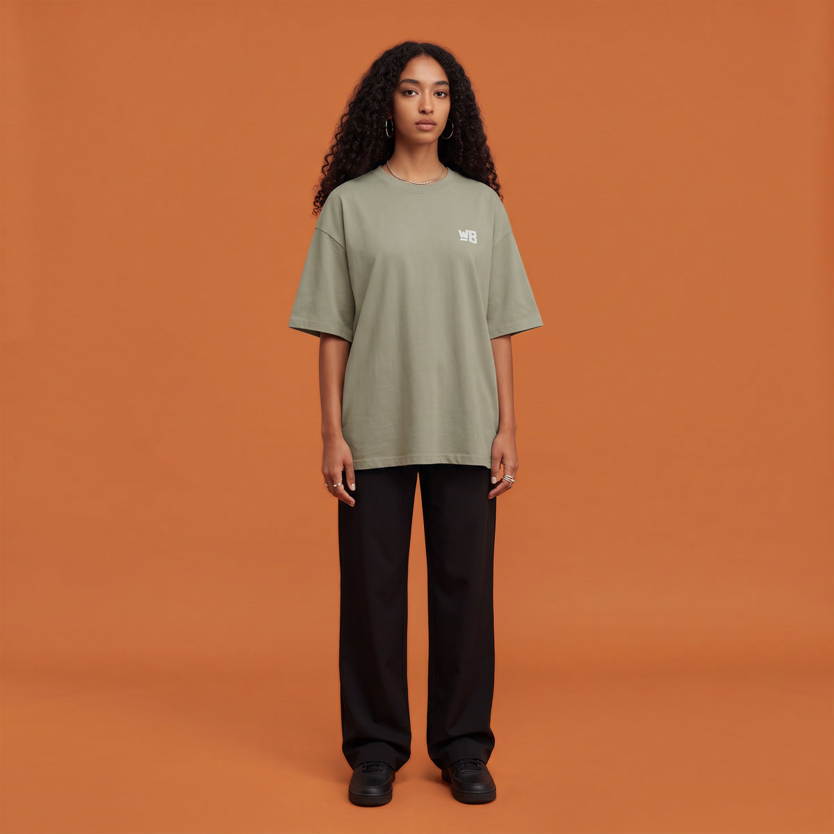 Blackness Solitude Tee, brown khaki, worn by a model — front view with WB chest logo, terracotta-orange studio backdrop